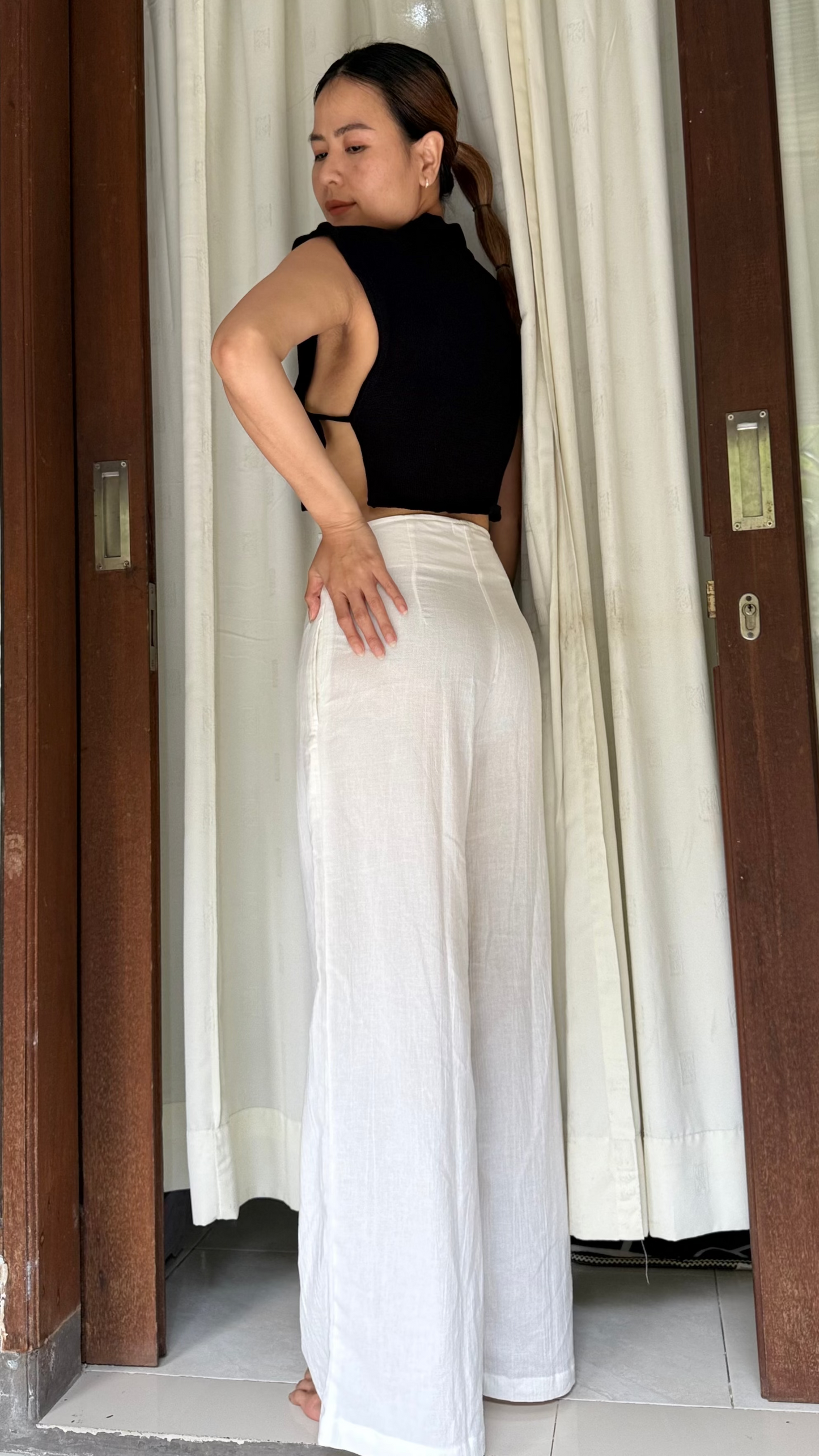 Woman in black top and white Lily pants standing in a doorway with curtains.